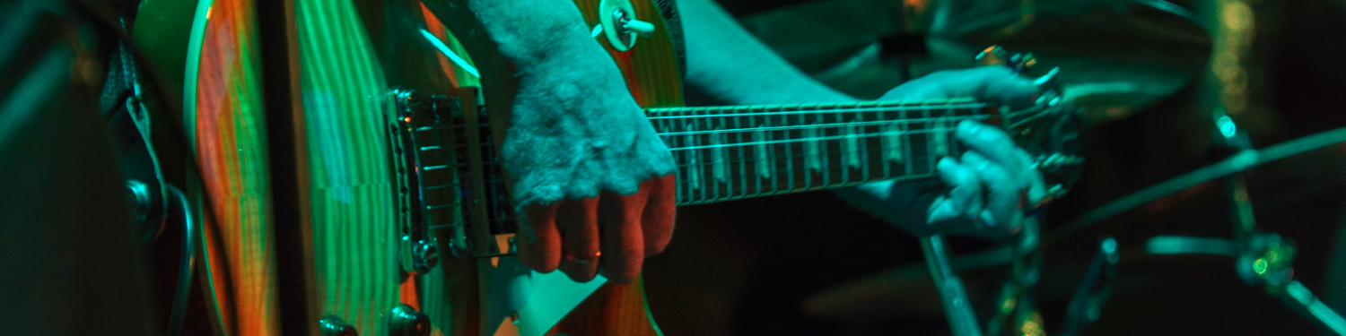 A close-up of a guitarist playing a wooden electric guitar, with hands on the neck and strings, vibrant stage lighting in greenish tones.