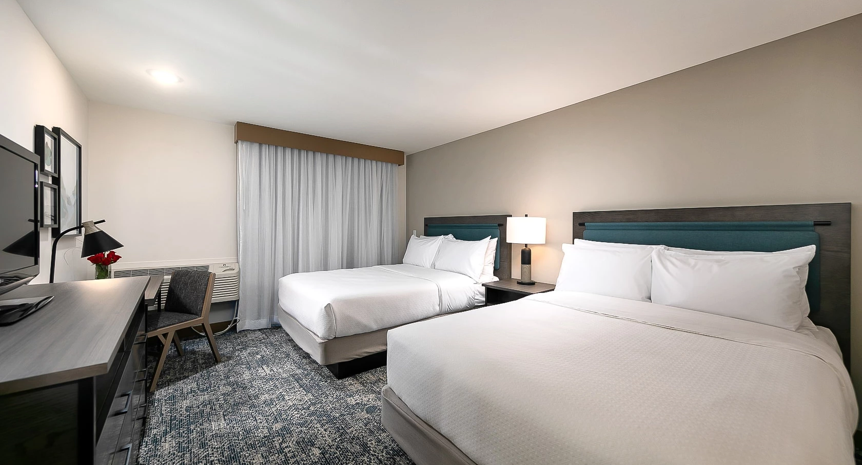 A clean hotel room with two queen beds, nightstand lamps, a work desk, chair, and a muted color scheme; window with curtains, patterned rug.