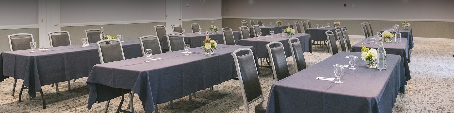 A conference room set for a meeting with multiple tables draped in dark blue cloths, glassware, and small floral centerpieces.