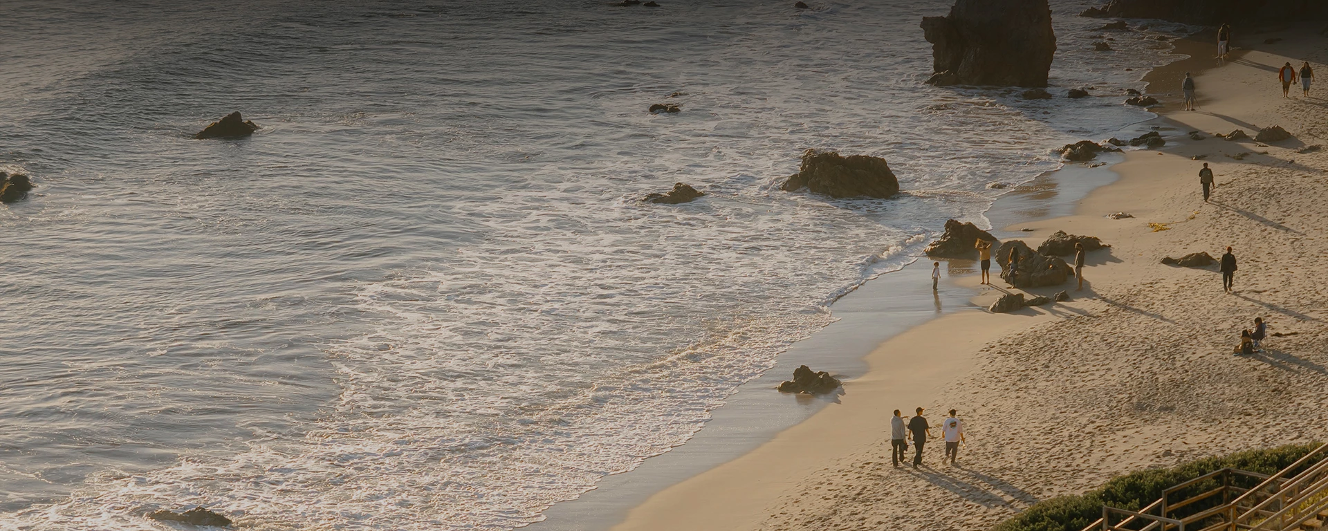 A sandy beach with gentle waves, a few groups strolling near the water, rocky outcrops, and warm evening light reflecting off the sea.