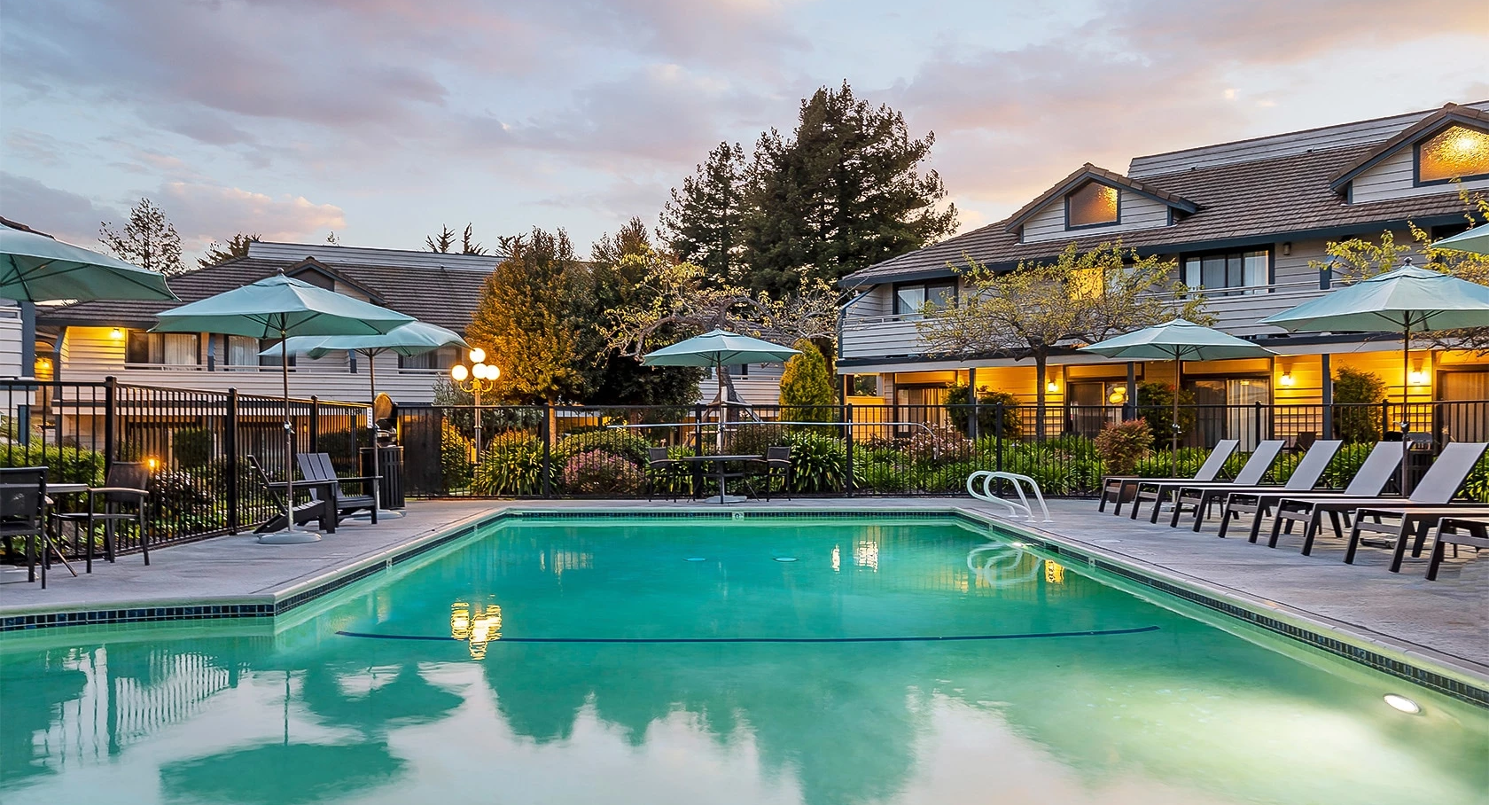 A calm hotel pool at dusk with lounging chairs, umbrellas, and warm lights reflecting off the water, surrounded by cozy buildings and trees.