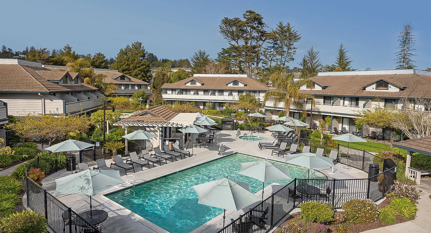 A sunny pool area with a fenced rectangular pool, lounge chairs under umbrellas, and tan multi-unit buildings around the courtyard.