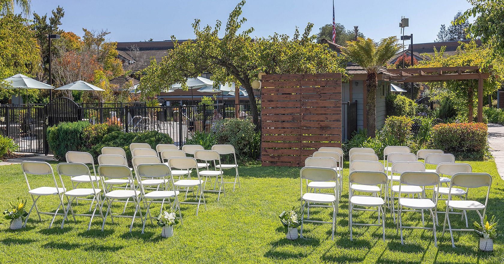 A grassy outdoor ceremony setup with white folding chairs arranged in rows, a wooden backdrop, and greenery under sunny skies.