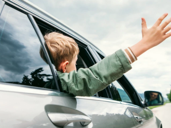 A child in a car window extends an arm out, smiling, as the car drives along a sunny, tree-lined road.