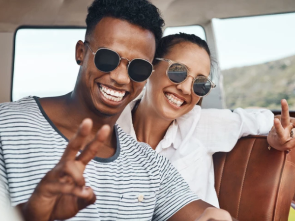 Two friends on a sunny day, smiling in a car, wearing sunglasses and making peace signs for a fun, casual selfie vibe.
