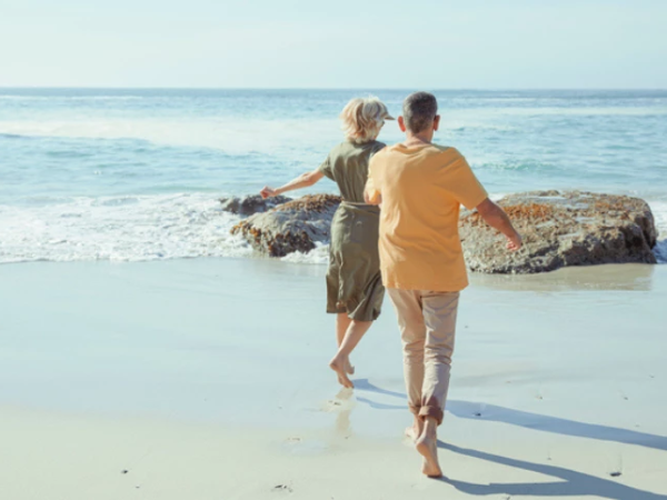 Two people walk hand-in-hand toward the ocean on a sunny beach, leaving footprints in the wet sand as waves roll in.