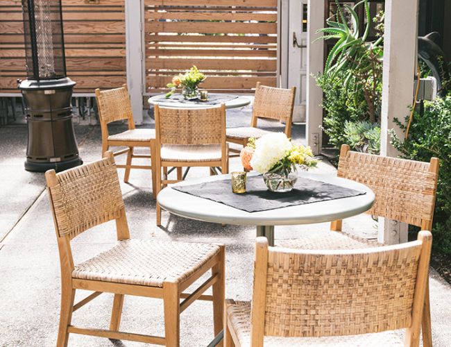 Outdoor patio with a round table and wicker chairs, potted plants, a tall heater, and a wooden privacy fence in a shaded cafe setting.