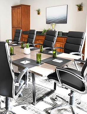 A modern conference room with a long glass table, 12 black ergonomic chairs, a wall TV, white walls, wood cabinets, plants, and large windows letting in light.