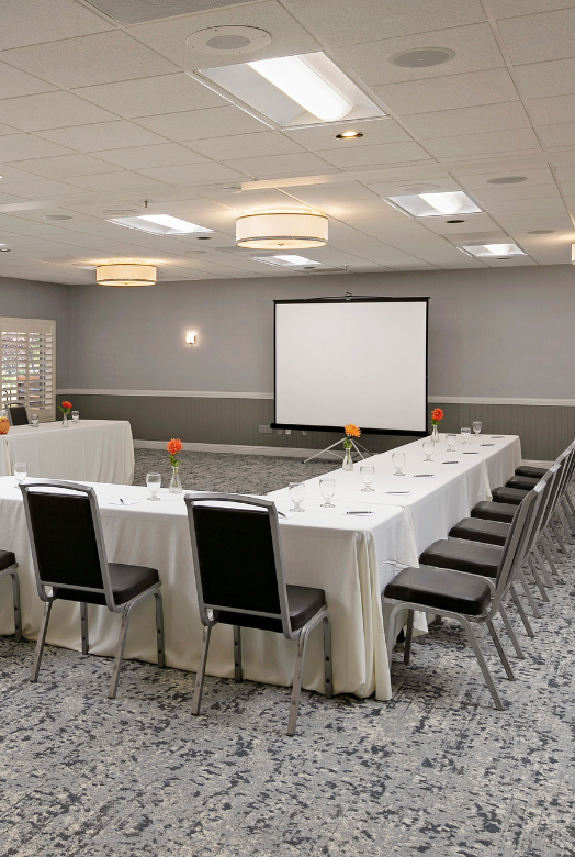 A large conference room with a U-shaped table setup, white tablecloths, black chairs, a projector screen, candles in bowls, and soft ceiling lighting.
