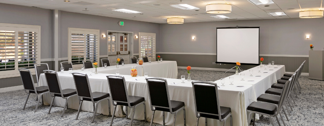 A large U-shaped conference setup with white tablecloths, black chairs, a projector screen, and neutral-toned banquet room decor.