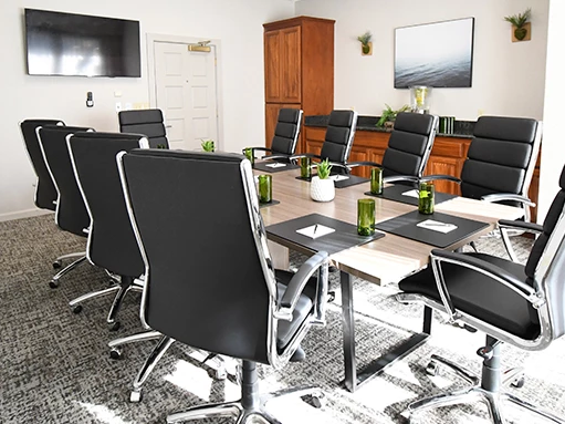 A modern conference room with a long glass table, black leather chairs, a wall-mounted TV, wooden cabinets, plants, and notebooks with green bottles on the table, sunny lighting.