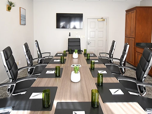 A modern boardroom with a long table, black leather chairs, green glass cups, white notepads, plants, and a wall TV.
