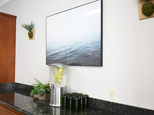 A kitchen or break room with a granite counter, a tall wooden cabinet, a blender with yellow liquid, framed ocean picture, small potted plants, and a row of empty bottles on the counter.