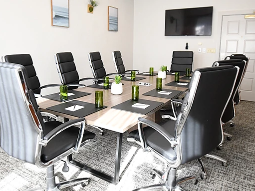 A modern conference room with a long table, black leather chairs, notebooks, green bottles, wall art, and a TV mounted on the wall.