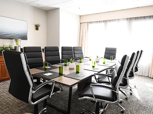 A modern conference room with a long table, black leather chairs, a wall-mounted TV, cabinets, plants, and green bottles set for a meeting.