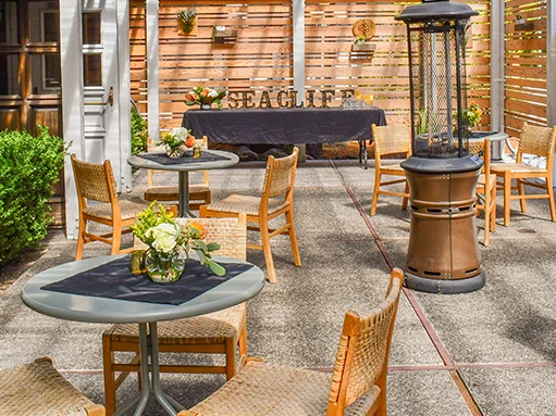 An outdoor seating area with wicker chairs around round and rectangular tables, potted plants, a tall patio heater, and a wooden fence backdrop.