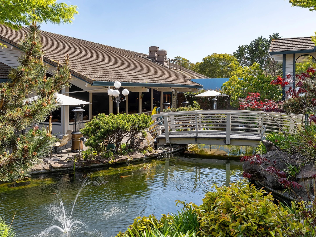 A serene outdoor dining area beside a canal, with a wooden bridge, greenery, and a building with a brown-tiled roof overlooking calm water.