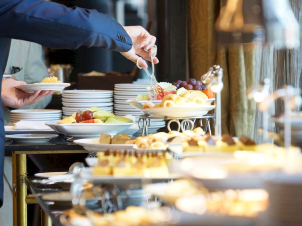 A person in a blue sleeve serves themselves at a buffet with plates, bowls, and a variety of desserts and fruit on a tiered display.