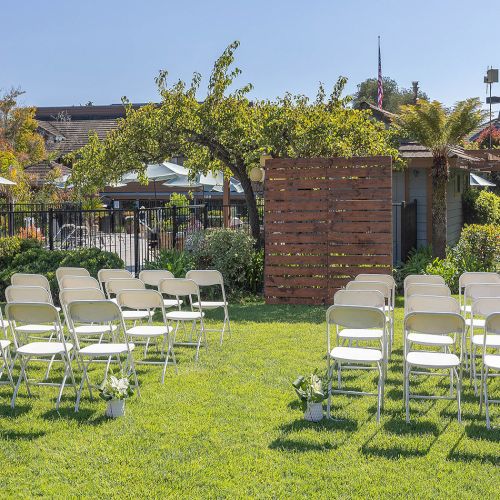 A daytime outdoor ceremony setup with two sections of white folding chairs on green grass, a wooden backdrop, plants, and shade structures in the background, under a clear sky.