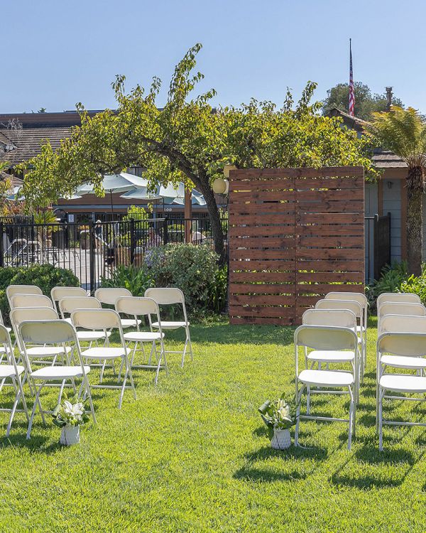 A daytime outdoor ceremony setup with two sections of white folding chairs on green grass, a wooden backdrop, plants, and shade structures in the background, under a clear sky.
