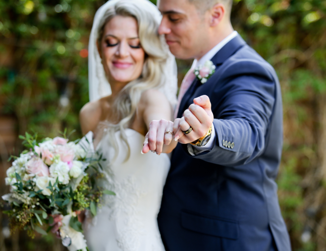A happy couple on their wedding day, the bride holds a bouquet while the groom shows off their rings, both smiling as they celebrate.