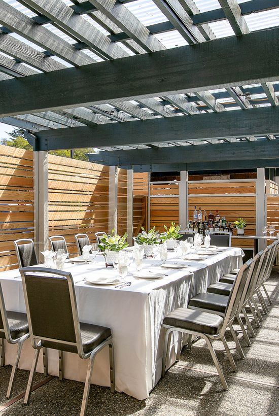 A long dining table set for a formal outdoor meal under a shaded pergola, with white linens, glassware, and metal chairs on a sunlit terrace.