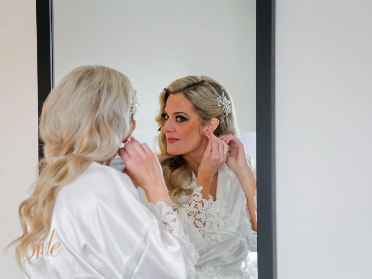 A bride in a lace robe adjusts her earring while looking at herself in a mirror.