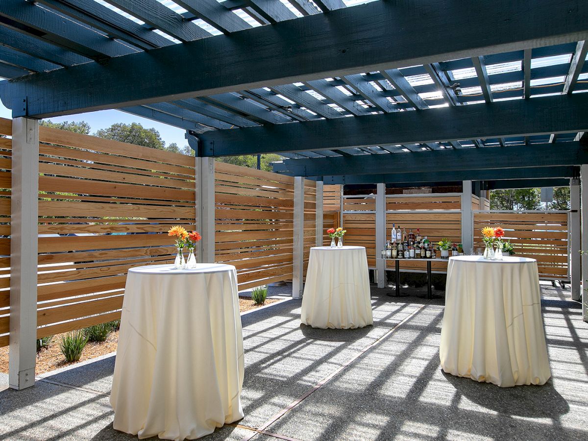 A shaded outdoor patio with a blue pergola, wooden privacy fences, and tall round tables draped in white cloths, set with small floral centerpieces.