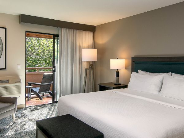 A modern hotel room with a king bed, two nightstands, a bench at the foot, lamps, and a sliding door opening to a balcony.
