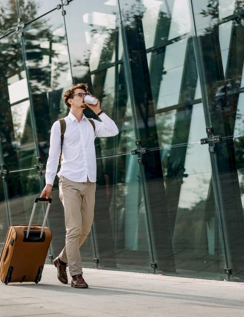 A person with a backpack walks outside a glass building, pulling a brown rolling suitcase and talking on a phone.