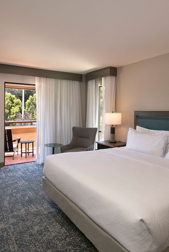 A modern hotel room with a king bed, teal headboard, two nightstands, lamps, a seating area near a balcony, and a wall-mounted TV.