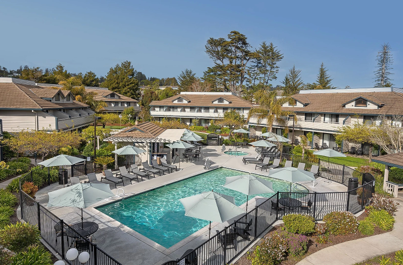 An outdoor hotel pool area with a rectangular pool, lounge chairs, umbrellas, and surrounding low-rise buildings in a sunny courtyard.