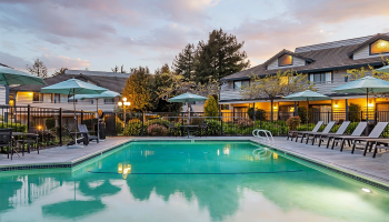 A serene hotel pool area at dusk with a turquoise pool, lounge chairs, umbrellas, and a two-story building in the background, warm lights glowing.