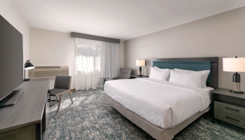A clean hotel room with two queen beds, matching nightstands, a desk and chair, a window with sheer curtains, and soft, neutral decor.