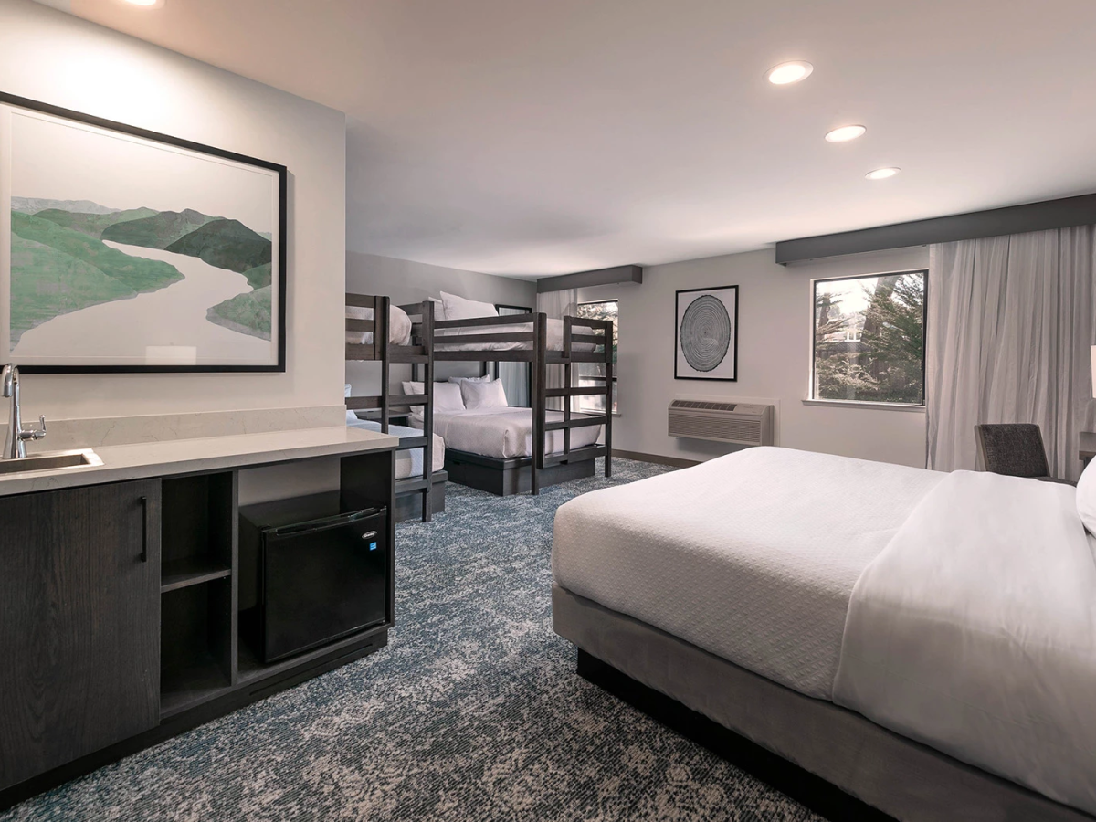 Modern hotel room with two double beds and a bunk bed setup, desk, mini-fridge, and a seating area by a window. It&rsquo;s clean, neutral-toned, and well-lit.