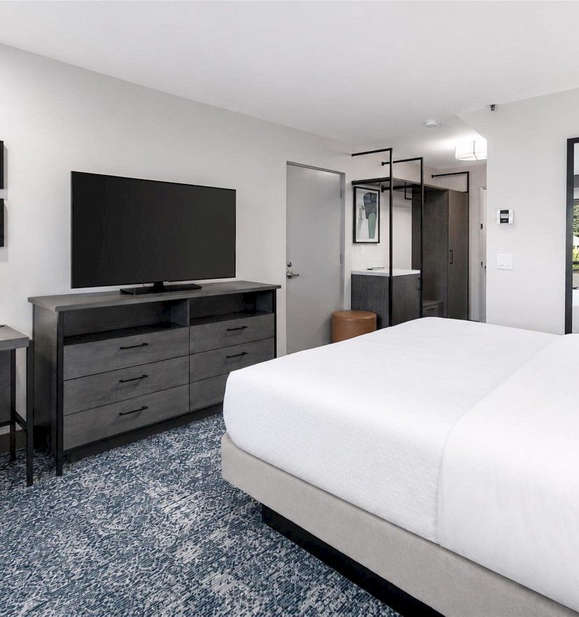 Modern hotel bedroom with a large white bed, flat-screen TV, dark dresser, framed wall art, and a blue patterned rug.