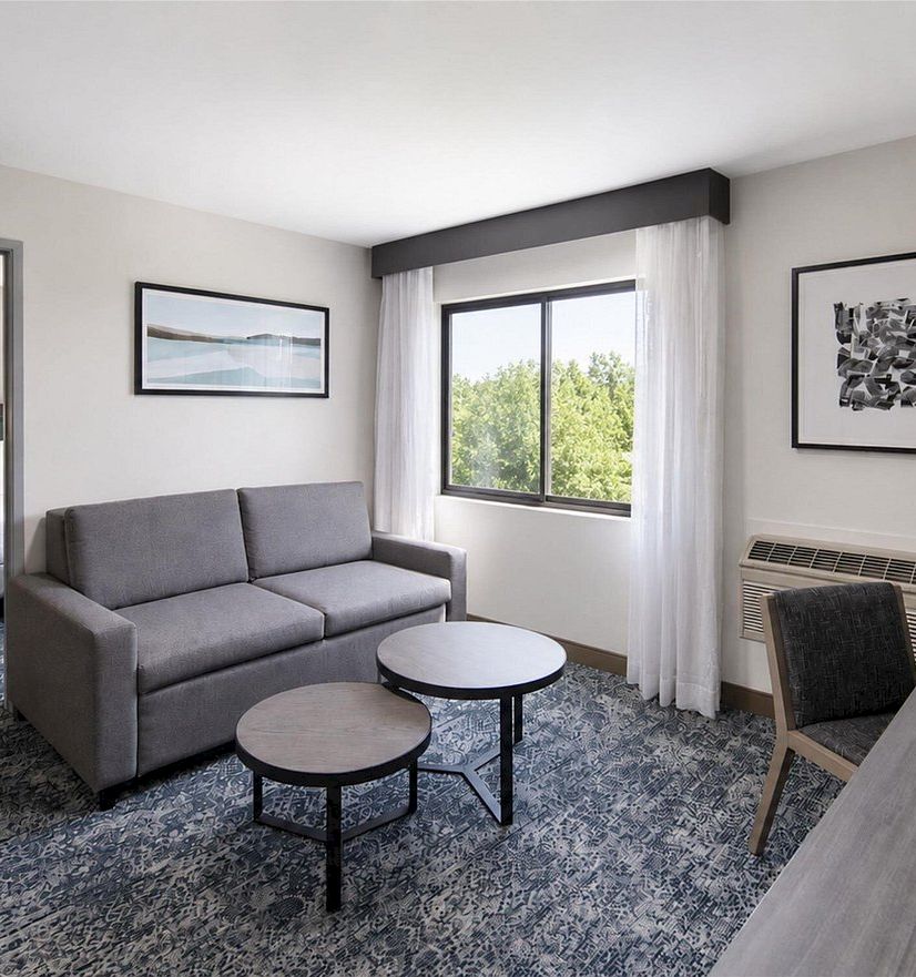 A modern hotel lounge with a gray sofa, round tables, framed art, curtains, a window view, and a wall AC unit.