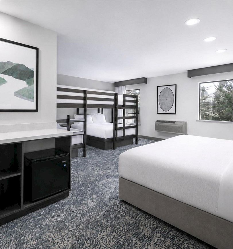 A modern hotel room with a large bed, minibar cabinet, sink/vanity, air conditioner, framed art, and tiled carpet.