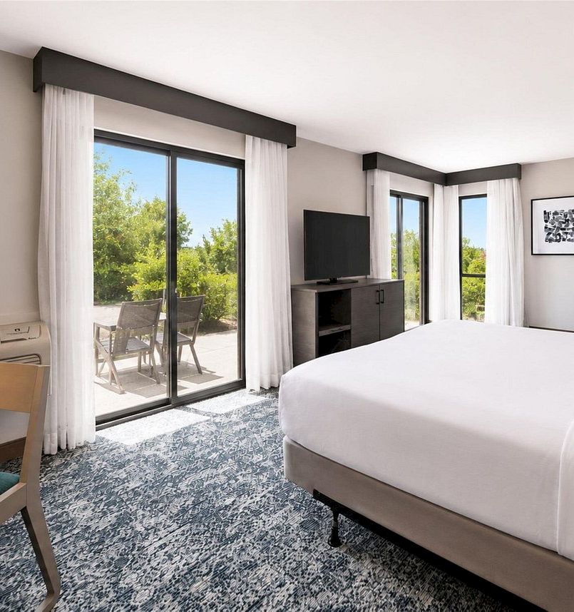 A modern hotel room with a white bed, TV stand, large windows, curtains, and an outdoor patio with chairs visible.