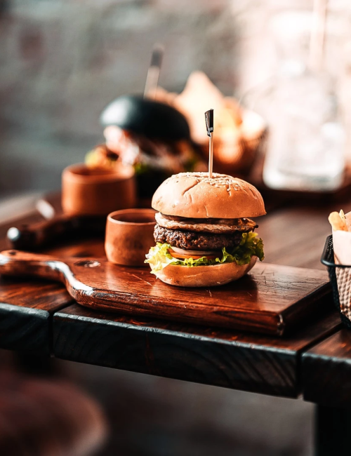 A cozy restaurant table with a burger, fries, and drinks on wooden boards and a dim, rustic backdrop.