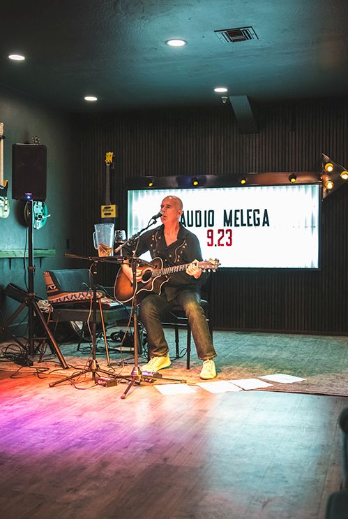 A guitarist performing on a small stage in a cozy music venue with amps, a screen, and audience chairs.