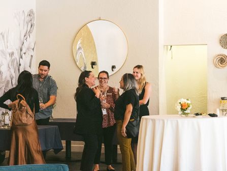 A group of people chat and mingle in a stylish, modern lounge with round mirror, white walls, and a high table setup; cozy seating and flowers.
