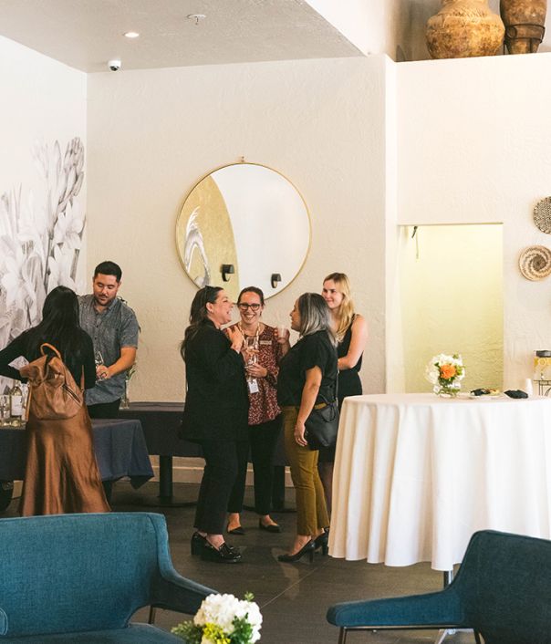 A group of people chat and mingle in a stylish, modern lounge with round mirror, white walls, and a high table setup; cozy seating and flowers.
