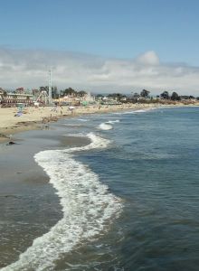 A sunny beach scene with people along the shore, soft waves rolling in, and buildings in the background by the coast.