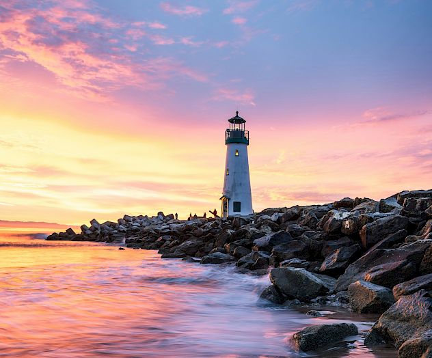 Coastal scene with a lighthouse on rocky shore at sunset, pink-orange sky reflecting on calm water, calm glow over waves and rocks.