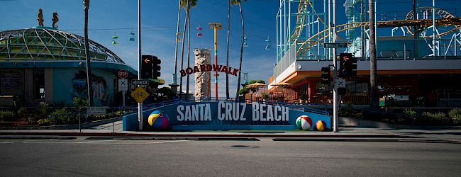 A sunny street scene at Santa Cruz Beach boardwalk with palm trees, a blue sign, and a tall roller coaster structure in the background.