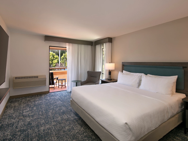 A modern hotel room with a large bed, two nightstands and lamps, a seating area by the window, and a balcony outside.