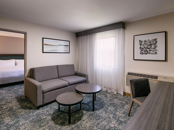 A modern hotel suite with a gray sofa, two round coffee tables, a desk and chair, framed art, a window with sheer curtains, and a separate bedroom in the background.