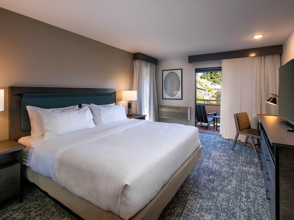 A modern hotel room with a large bed, two nightstands, a desk and chair, a wall-mounted TV, and a balcony visible through sliding doors, all in neutral tones.