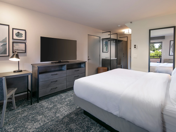 A modern hotel room with a king bed, desk, dresser, wall art, flat-screen TV, and a glass-enclosed shower area in the background.